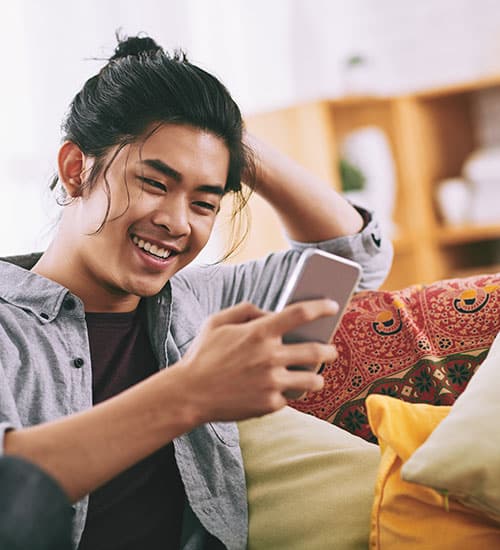 Young person sitting on a couch, smiling while looking at a smartphone, with one hand behind their head and colorful pillows in the background.