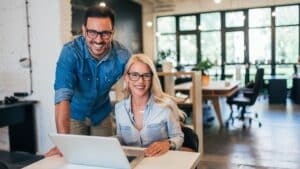 Two people wearing glasses and denim shirts are smiling at a desk with a laptop in a modern, well-lit office space.