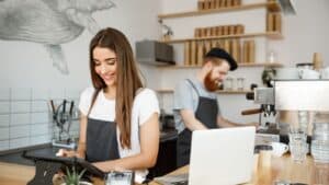 Two baristas wearing aprons work in a modern café; one uses a tablet at the counter, while the other prepares drinks near a coffee machine in the background.